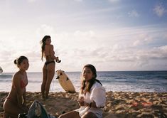 three women sitting on the beach with surfboards in front of them and one woman standing next to her