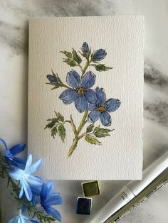 some blue flowers are sitting on a table next to markers and paintbrushes with watercolor pencils