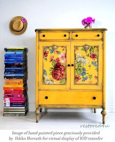 a yellow cabinet with flowers painted on it next to a stack of books and a hat