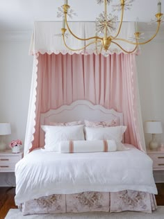 a white bed with pink curtains and pillows in a bedroom next to a chandelier