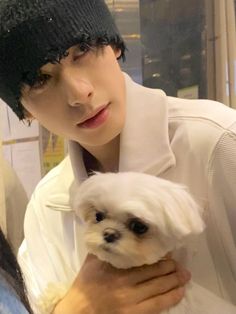 a man holding a small white dog in his arms while wearing a beanie hat