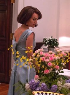 a woman in a blue dress standing next to a vase with flowers on the table