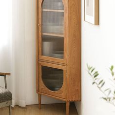 a tall wooden cabinet with glass doors next to a chair and potted plant on the floor