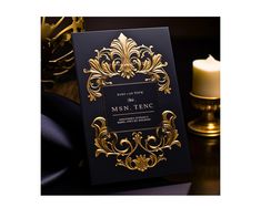 a black and gold wedding card with an ornate design on the front, sitting next to a candle