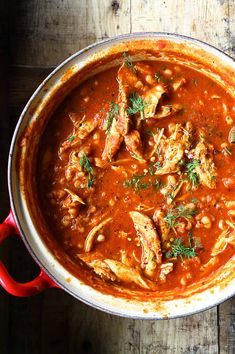 a red pot filled with stew and meats on top of a wooden table next to a spoon