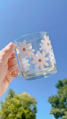 a hand holding a clear glass with white daisies on the side and trees in the background