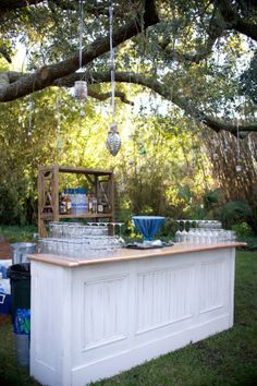 an outdoor bar is set up under a tree with wine glasses on it and bottles in the foreground