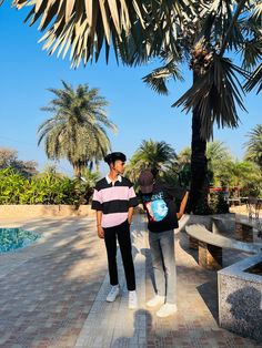 two men standing next to each other on a brick walkway near a palm tree and pool