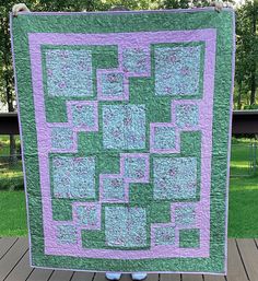 a large green and purple quilt on a wooden deck with trees in the back ground