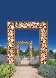 an arch made out of logs in the middle of a garden with blue sky and lavenders