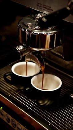 coffee being poured into two white cups in front of an espresso machine with steam coming from it