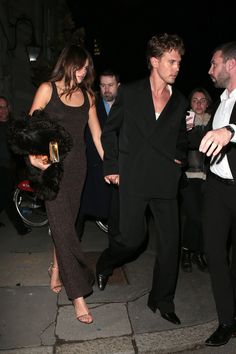 a man and woman walking down the street at night with other people in the background