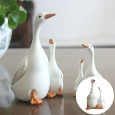 three white ducks standing next to each other on top of a wooden table with a potted plant in the background