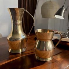 two shiny metal pitchers sitting on top of a wooden table next to a desk lamp