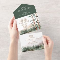 two hands holding up a green and white wedding card with trees in the woods on it