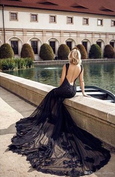 a woman in a black dress is sitting on a ledge looking at the water and buildings