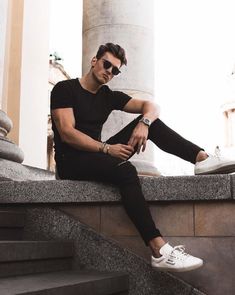 a man sitting on some steps with his legs crossed and wearing white tennis shoes, black t - shirt and jeans