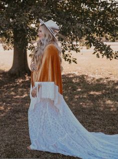 a woman in a white dress and hat is standing under a tree with her back to the camera