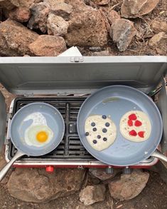 two pans with food cooking on top of them in front of rocks and gravel