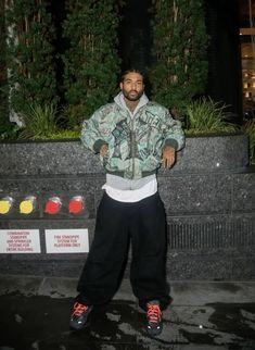 a man in camouflage jacket and black pants standing next to a wall with plants on it