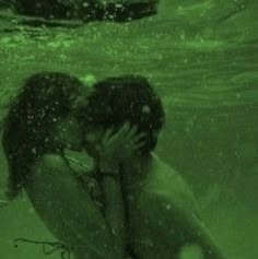 two people kissing in the water with their arms around each other, under green light