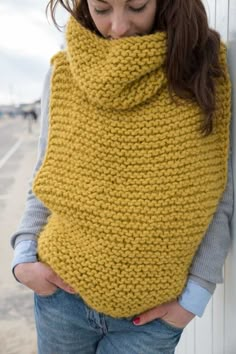 a woman wearing a yellow knitted cowl with her hands on her hips, leaning against a wall