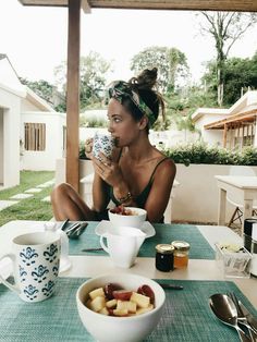 a woman sitting at a table drinking from a cup with fruit in front of her