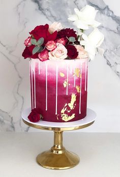 a red and white cake with flowers on top is sitting on a gold stand in front of a marble wall