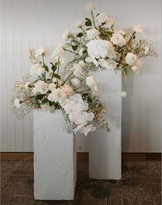 two white vases with flowers in them sitting on a carpeted floor next to a wall