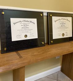 two framed diplomas sitting on top of a wooden table in front of a wall