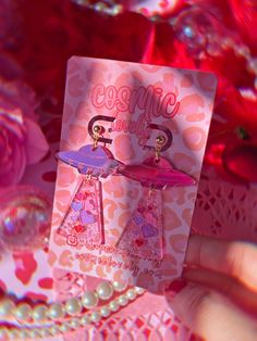 a pair of pink earrings is being held up by someone's hand in front of some flowers