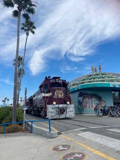 a train passing under a bridge with palm trees in the foreground and people on bicycles behind it