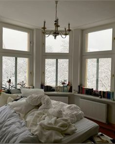 an unmade bed in front of three windows with books on the shelves and a chandelier hanging from the ceiling