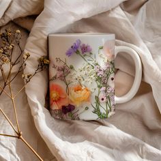 a coffee mug with flowers painted on it sitting on a white blanket next to dried plants