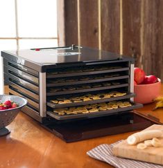 An L.E.M. brushed nickel food dehydrator sits on a table in front of a window. A fresh banana sits on a cutting board in the foreground. There are banana slices on each rack of the dehydrator. Homemade Trail Mix, Crunchy Granola, Fruit Roll, Fruit Puree, Homemade Yogurt, Digital Timer