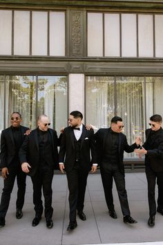 four men in tuxedos are standing near a building and one man is holding his arm around another man's shoulder