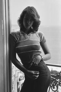 a black and white photo of a woman leaning against a window sill with her hands on her hips