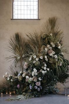 an arrangement of flowers and greenery in front of a window with candles on the floor