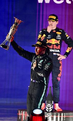 two men on stage holding up trophies and wearing black outfits with red lettering behind them