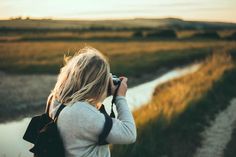 a woman with a camera is standing in front of a river and looking at the grass