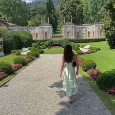 a woman walking down a path in front of a garden with hedges and flowers on both sides