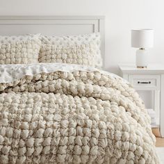 a white bed with two nightstands in the background and a large chunky blanket on top of it