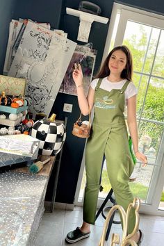 a woman in overalls standing next to a table with art on the wall behind her