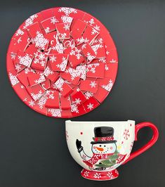 a red and white coffee cup sitting next to a plate with snowflakes on it