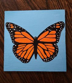 an orange and black butterfly painted on a blue square with white polka dot dots, sitting on a wooden surface