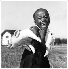 a woman holding a baby pig in her arms while standing in a field with a house in the background