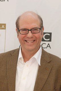 an older man wearing glasses and a brown jacket smiles at the camera while standing in front of a white wall