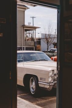 an old white car is parked in a parking lot near a building with a clock on it's side