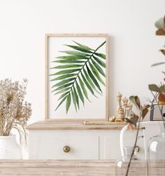 a green palm leaf in a wooden frame on a white wall above a table with vases and plants