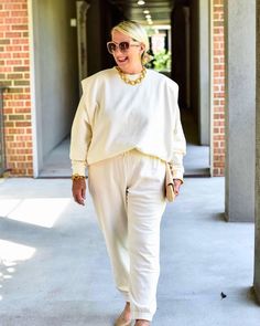 an older woman walking down the sidewalk wearing white pants and a cream sweater with gold necklace